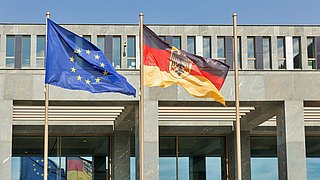 Foto, eine Europa- sowie eine Deutschlandflagge wehen vor einem Gebäude.